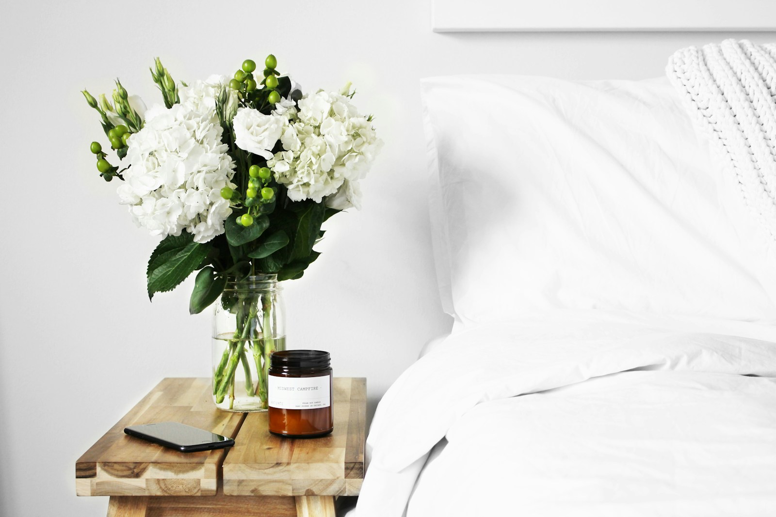 flowers in clear glass vase on bedside table next to bed with white bedding